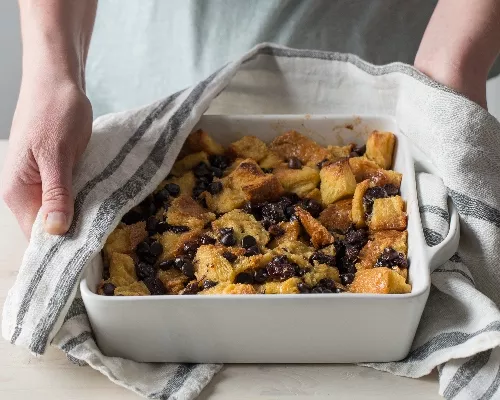 Challah Bread Pudding with Chocolate and Dried Fruit in a serving dish being held with a dish towel