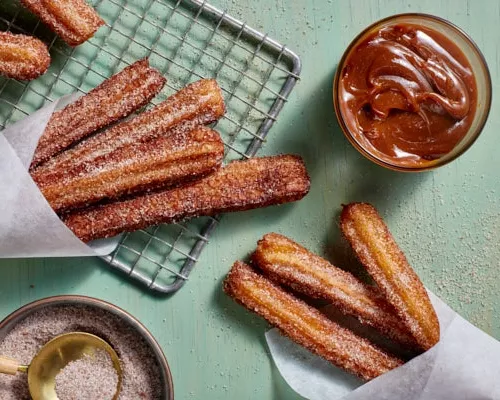   Churros with cinnamon sugar on a wire cooling rack with a bowl of dulce de leche dip