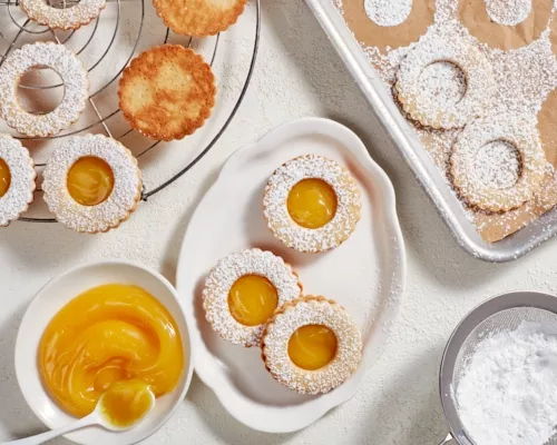 Citrus curd cookies on a platter shown with unfinished cookies on a cooling rack and a baking pan and a bowl of citrus curd