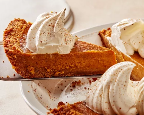 Removing a slice of pumpkin pie with whipped cream and ground cinnamon from a white pie plate with a pie lifter.  