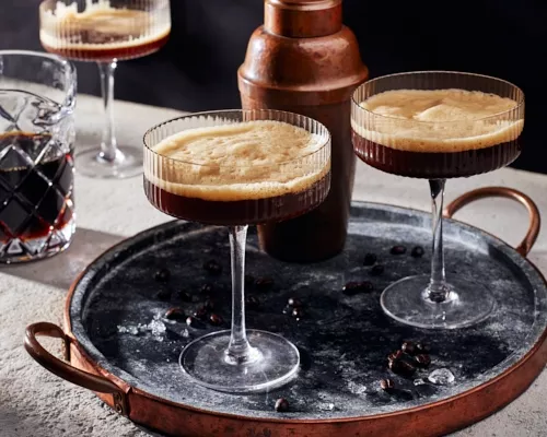 Two glasses of frothy espresso martini on a tray with a cocktail shaker with a third glass and a glass pitcher of espresso marti