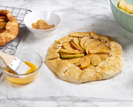 Unbaked apple galette sprinkled with turbinado sugar, and egg wash with a brush in a bowl