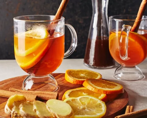 Two hot toddies in glass mugs garnished with orange slices and cinnamon sticks, one on a wood platter with garnishes and sliced
