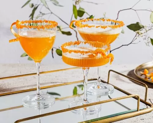 Three glasses of sparkling spiced ginger turmeric cocktail garnished with orange peel and rimmed with sugar on a mirrored tray