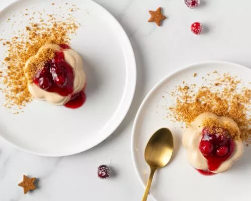 Two gingerbread panna cotta topped with cranberries on white plates with a gold spoon