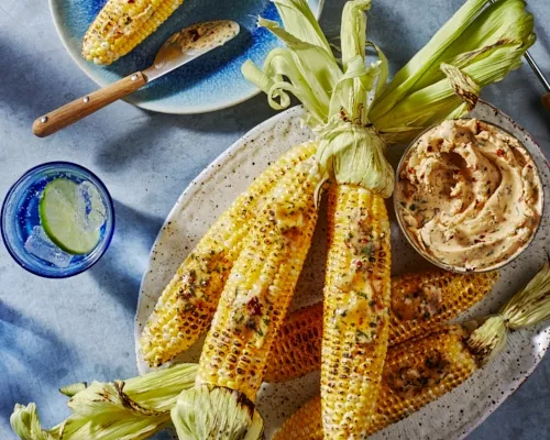  Grilled corn on the cob on a platter with a bowl of chipotle-lime butter, shown with a glass of ice water with a slice of lime