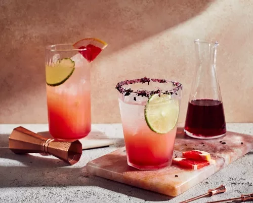 Two glasses of hibiscus paloma with a pitcher of simple syrup, shown with a jigger and skewers.