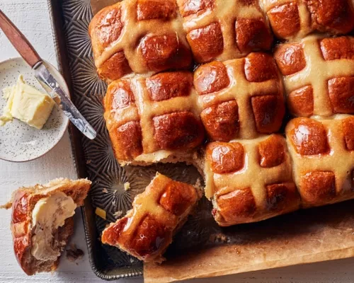 Hot cross buns on a baking sheet with one removed, sliced, and buttered