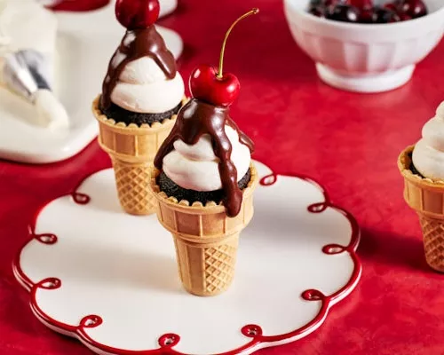 Chocolate cupcakes in classic ice cream cones decorated with icing, chocolate ganache, and topped with a cherry served on a red 