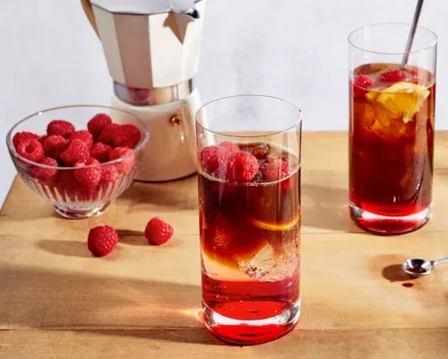 Two glasses of iced raspberry espresso tonic on a wooden table shown with a bowl of raspberries and an espresso maker