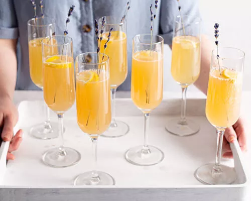 Seven champagne flutes of mimosa on a serving tray held by two hands