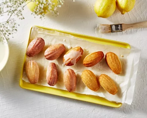 Glazed lemon madeleines on a yellow tray shown with a bowl of glaze, a glazing brush, and lemons