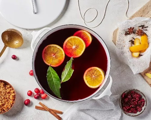 A pot of mulled wine with orange slices and bay leaves shown with sliced ginger, cranberries, and brown sugar