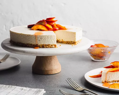 No-bake vanilla cheesecake with peach topping on a cake stand with a slice served on a plate.