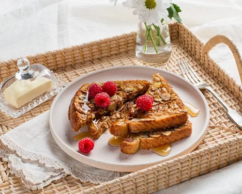 Oat and almond-crusted French toast on a rattan serving tray with butter in a glass dome and flowers in a vase