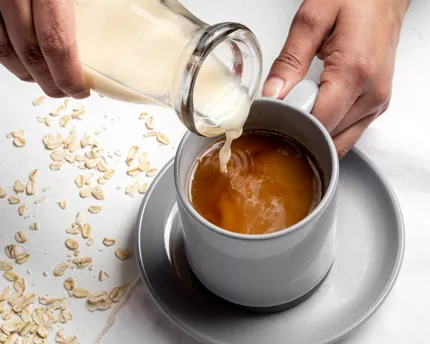 Pouring vanilla oat beverage into a mug of coffee from a glass bottle