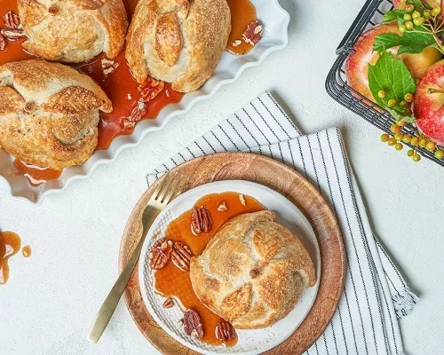 Three apple dumplings on a tray with apple cider syrup, and one on a plate