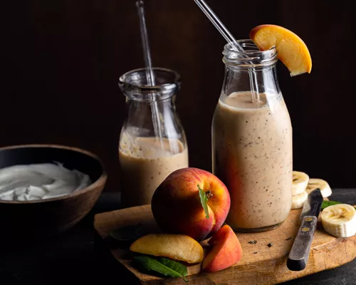 Two single serving bottles of banana peach Earl Grey smoothie with glass straws, with a cutting board and fruit