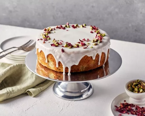 Persian Love Cake with icing and rose petals on a silver cake stand, shown with plates, forks, and napkins