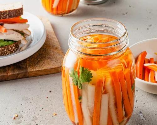 A jar of pickled carrots and daikon radishes with a sandwich in the background