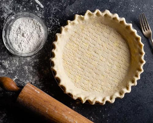 Unbaked pie crust with a bowl of flour, a rolling pin, and a fork