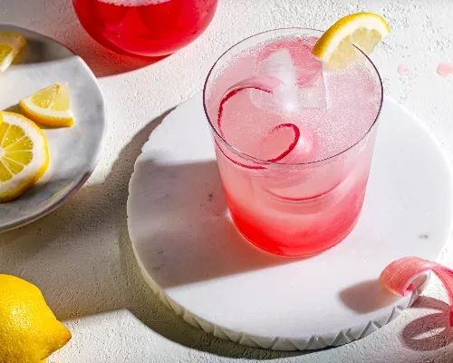 Glass of pink rhubarb lemonade on a coaster garnished with lemon and rhubarb, and a plate of lemon wheels
