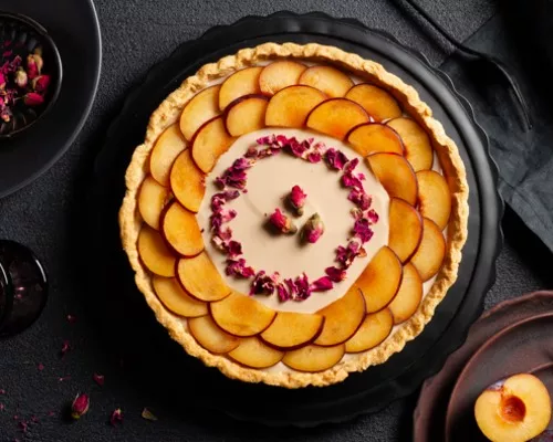 Plum custard tart decorated with sliced plums and dried rose petals on a black platter on a black table