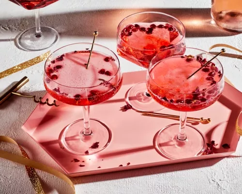 Three pomegranate-rose sparkling cocktails in stemmed glasses on a pink tray, shown with gold swizzles and gold ribbons