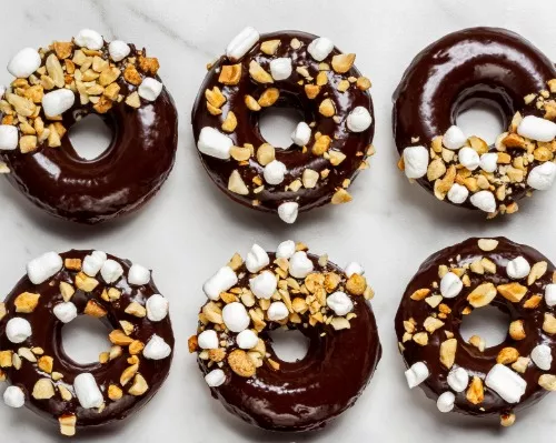 A chocolate donut with chocolate icing half sprinkled with peanuts and marshmallows with a bite missing