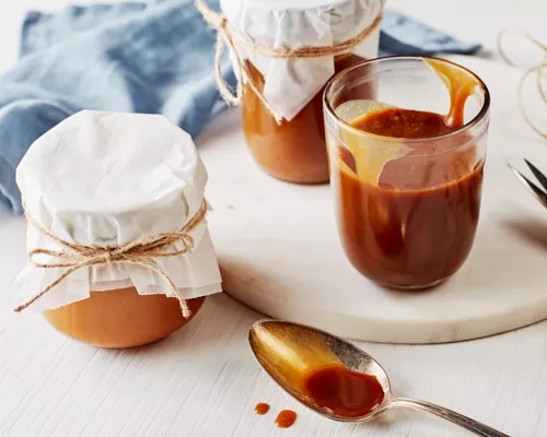 Three glass jars of salted butterscotch sauce, one open and two covered with cloth and tied off with twine.