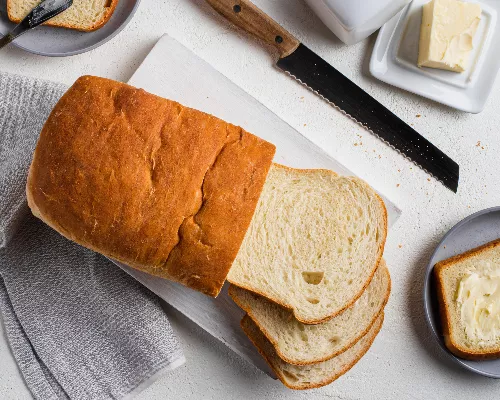 A partially sliced loaf of bread on a cutting board, with a bread knife and one slice buttered on a plate