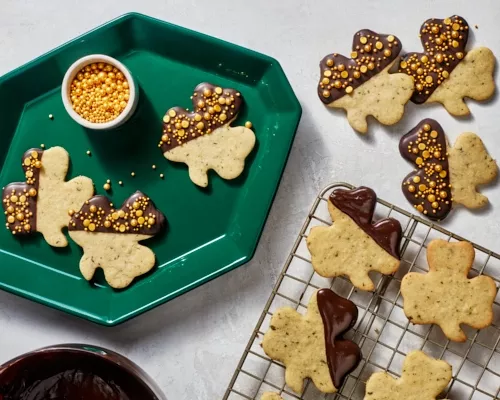 Shamrock-shaped mint cookies dipped in chocolate and decorated with gold sprinkles on a green tray and a wire cooling rack