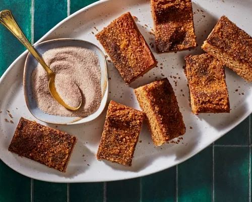 Seven snickerdoodle cookie bars on a white platter with a dish of cinnamon sugar and a gold spoon, shown on a green tile counter