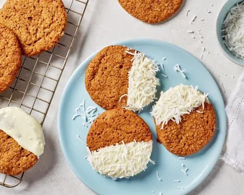 Gingersnap cookies half dipped in white chocolate and coconut on a blue plate and a wire cooling rack