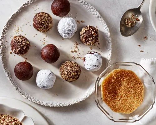 Spiced bourbon balls on a platter, topped with icing sugar, crushed nuts, and turbinado sugar, shown with bowls of toppings.