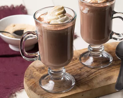 Two mugs of hot chocolate with whipped cream on top, sitting on a wooden board