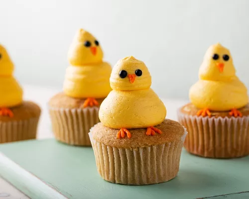 Cupcakes decorated with yellow chicks made of piped icing, on a green wood board