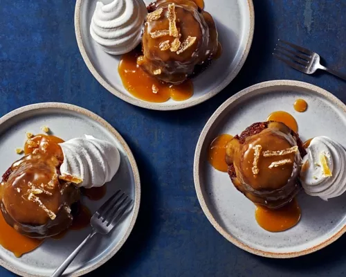 Top down view of three sticky toffee puddings on plates, each with a dollop of whipped cream and toffee sauce.