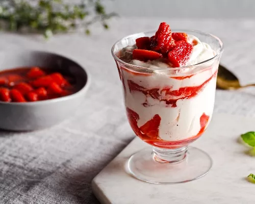 Strawberry fool in a parfait glass on a marble slab with a bowl of strawberries in sauce