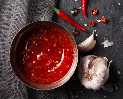 Metal bowl of sweet chili sauce with whole and cut chilis and a clove of garlic