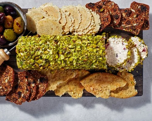 A cream cheese log coated in crushed pistachios on a serving board, surrounded by crisps and crackers and a bowl of olives