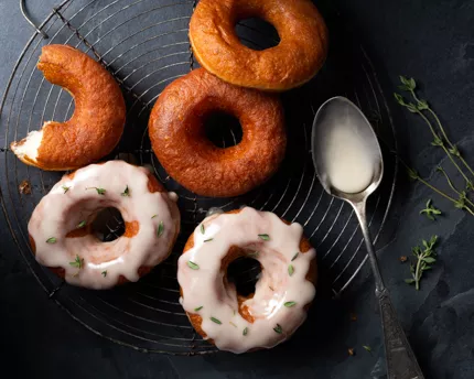 Five yeasted doughnuts on a cooling rack, two iced and sprinkled with thyme, with a spoon and thyme sprigs