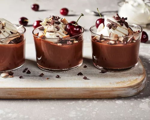 Three vegan chocolate puddings topped with whipped cream, coconut flakes, cacao nibs, and cherries, served on a cutting board