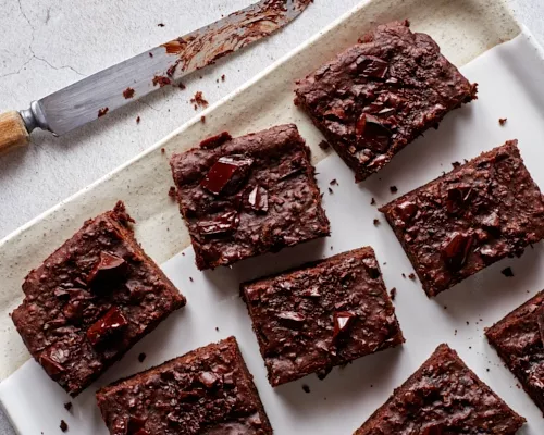 Vegan brownies topped with dark chocolate chunks on a platter with a knife.