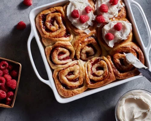 Fresh vegan raspberry cinnamon rolls in a baking dish, some bare, some with coconut glaze and raspberries