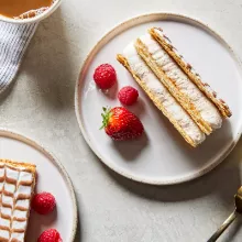 Two pieces of Mille-Feuille cake on plates, shown with berries, a cup of coffee and a gold fork.
