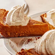 Removing a slice of pumpkin pie with whipped cream and ground cinnamon from a white pie plate with a pie lifter