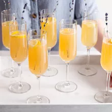 Seven champagne flutes of mimosa on a serving tray held by two hands