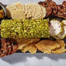 A cream cheese log coated in crushed pistachios on a serving board, surrounded by crisps and crackers and a bowl of olives