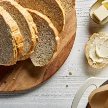 Un pain de levain sucré coupé en tranches sur une planche à découper en bois, avec une tranche beurrée, présenté avec un plat de beurre et un couteau à beurre.
